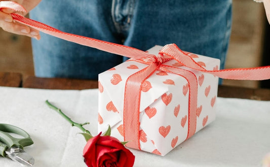 A person wrapping an Eco-friendly Valentine's Day gift in white paper with red hearts and tying a red ribbon